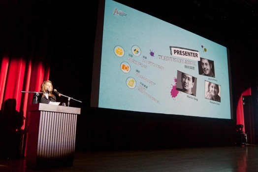 image of a woman on a stand and next to her with a big picture projected on the wall with a few headshots and some other text
