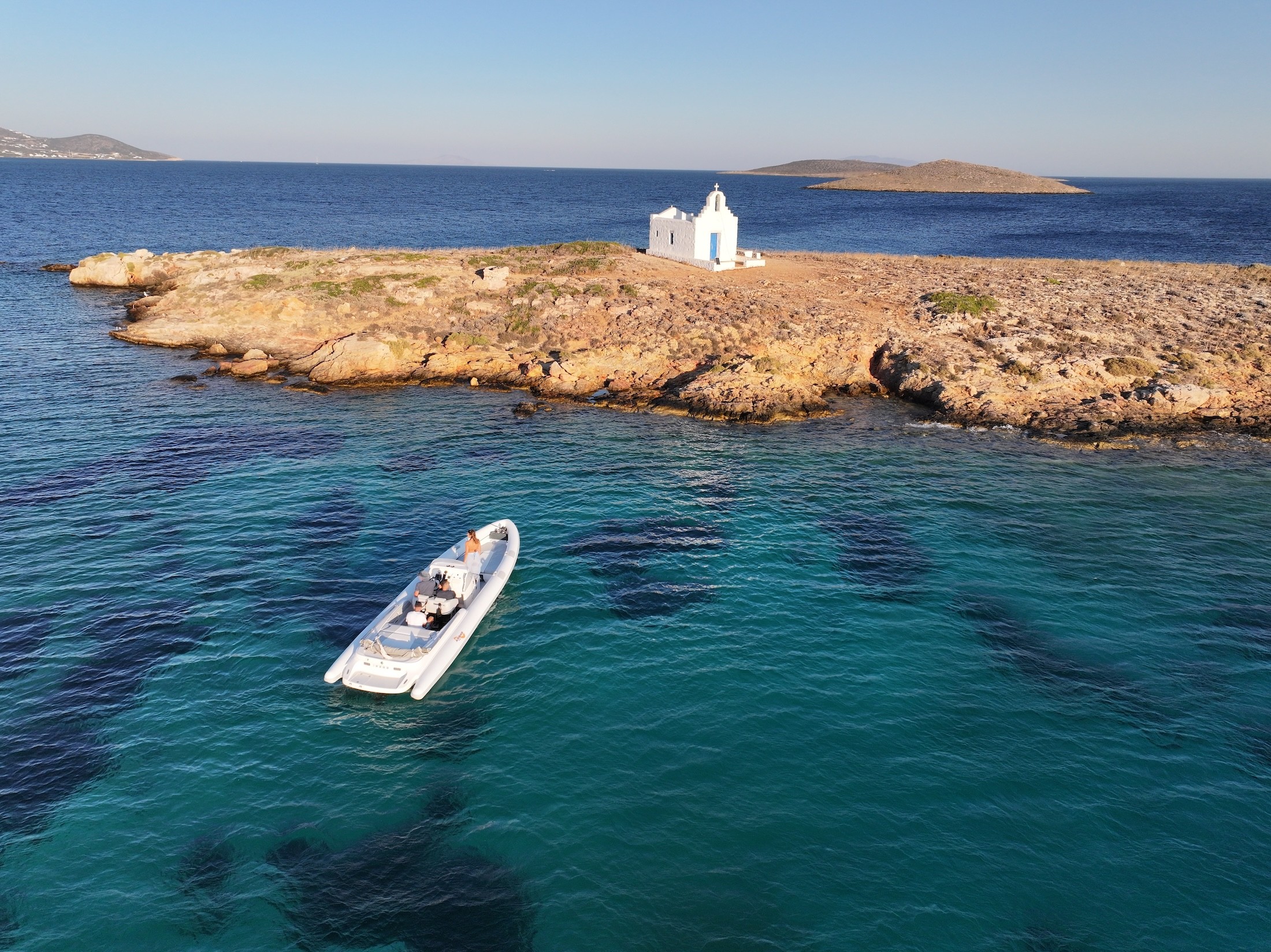 White Airship 30 Aether yacht anchored in crystal-clear turquoise waters off a rocky Cycladic coastline with traditional white houses.