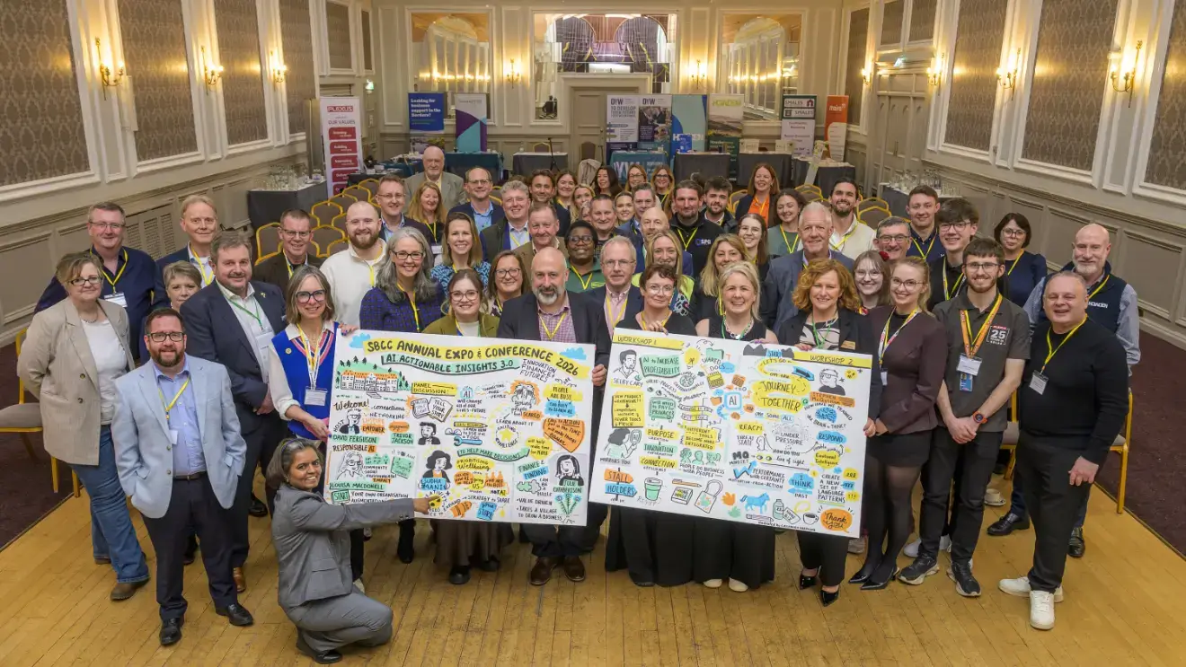 A large group of people at a conference smile for a photo, holding two illustrated posters. The room is elegantly decorated, exuding a warm, welcoming atmosphere.