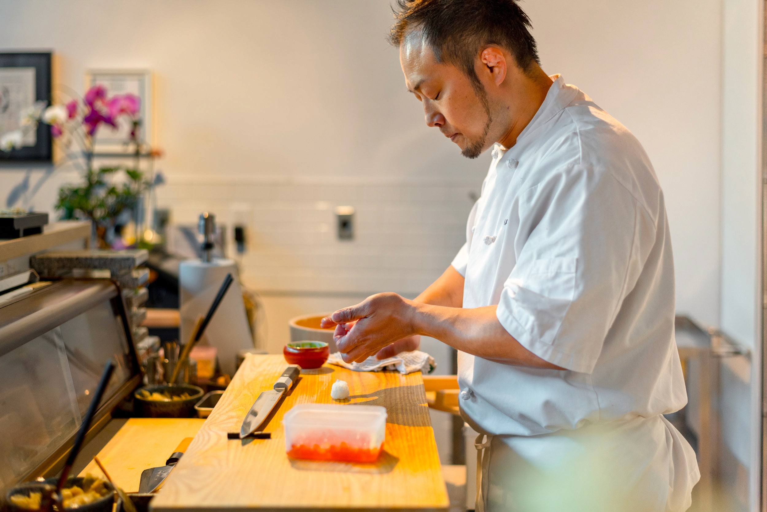 A sushi chef in a white uniform prepares sushi on a wooden counter in a brightly lit restaurant.