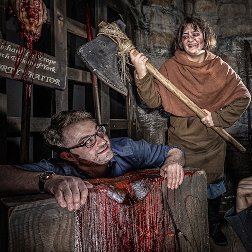 A man is locked in a medieval stock while a woman, smiling, raises an axe above him. Blood drips from the stock.