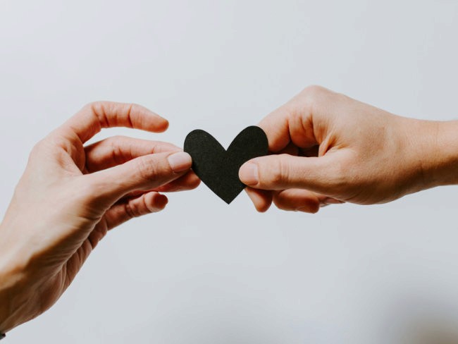 Zwei Hände halten gemeinsam ein kleines schwarzes Papierherz vor hellem Hintergrund