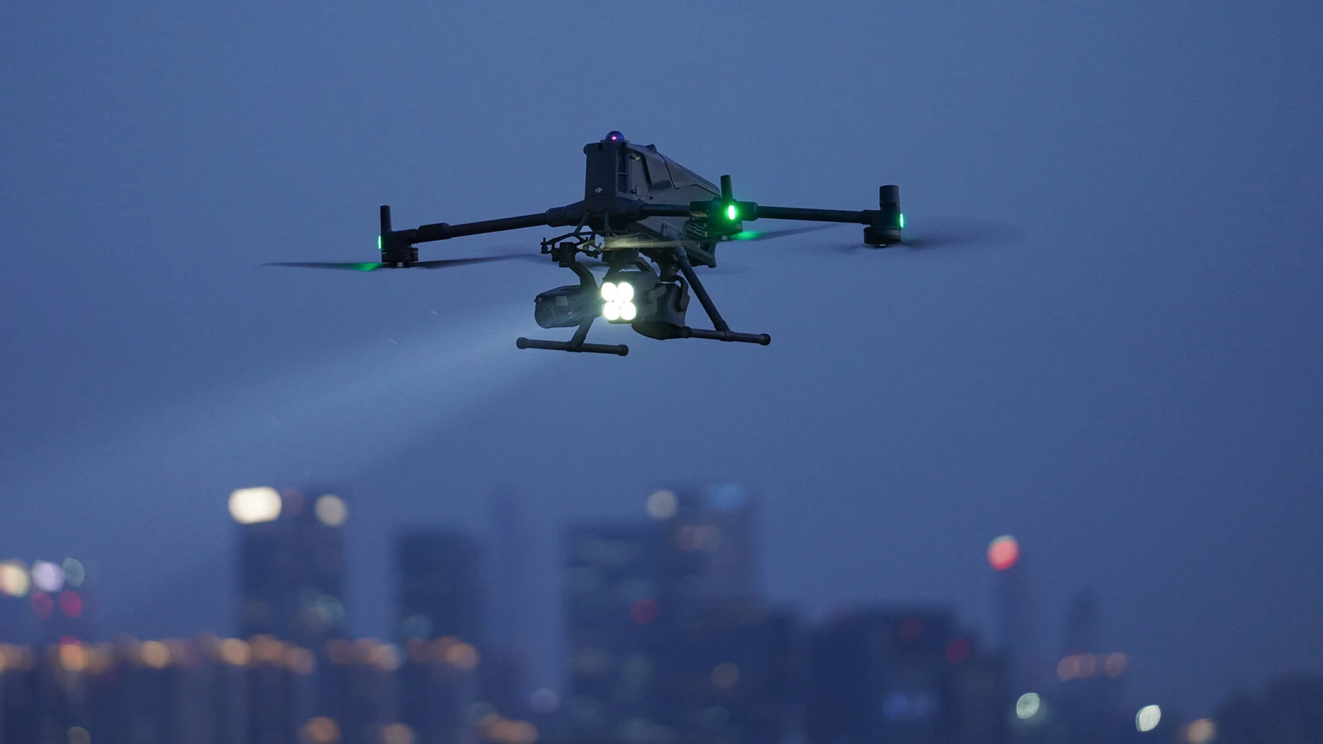 Drone performing night imaging inspection with spotlight over a city skyline after dark
