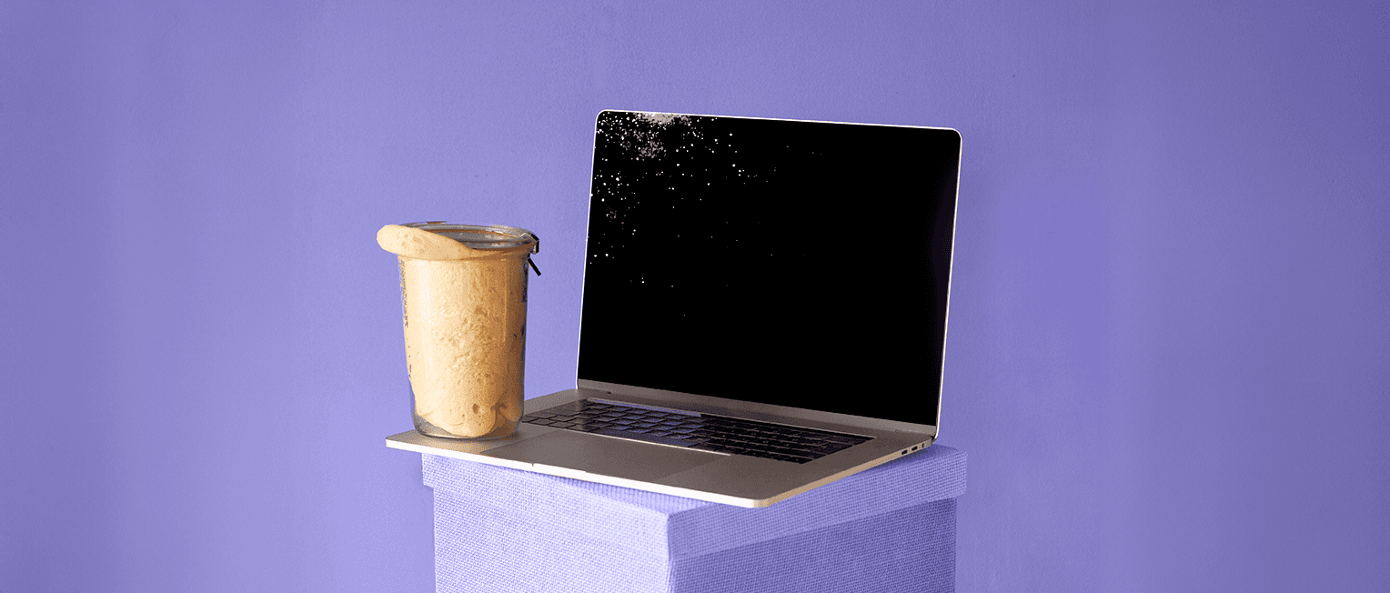 Laptop placed on a lila box, next to a jar with sourdough almost exiting it, on a purple background