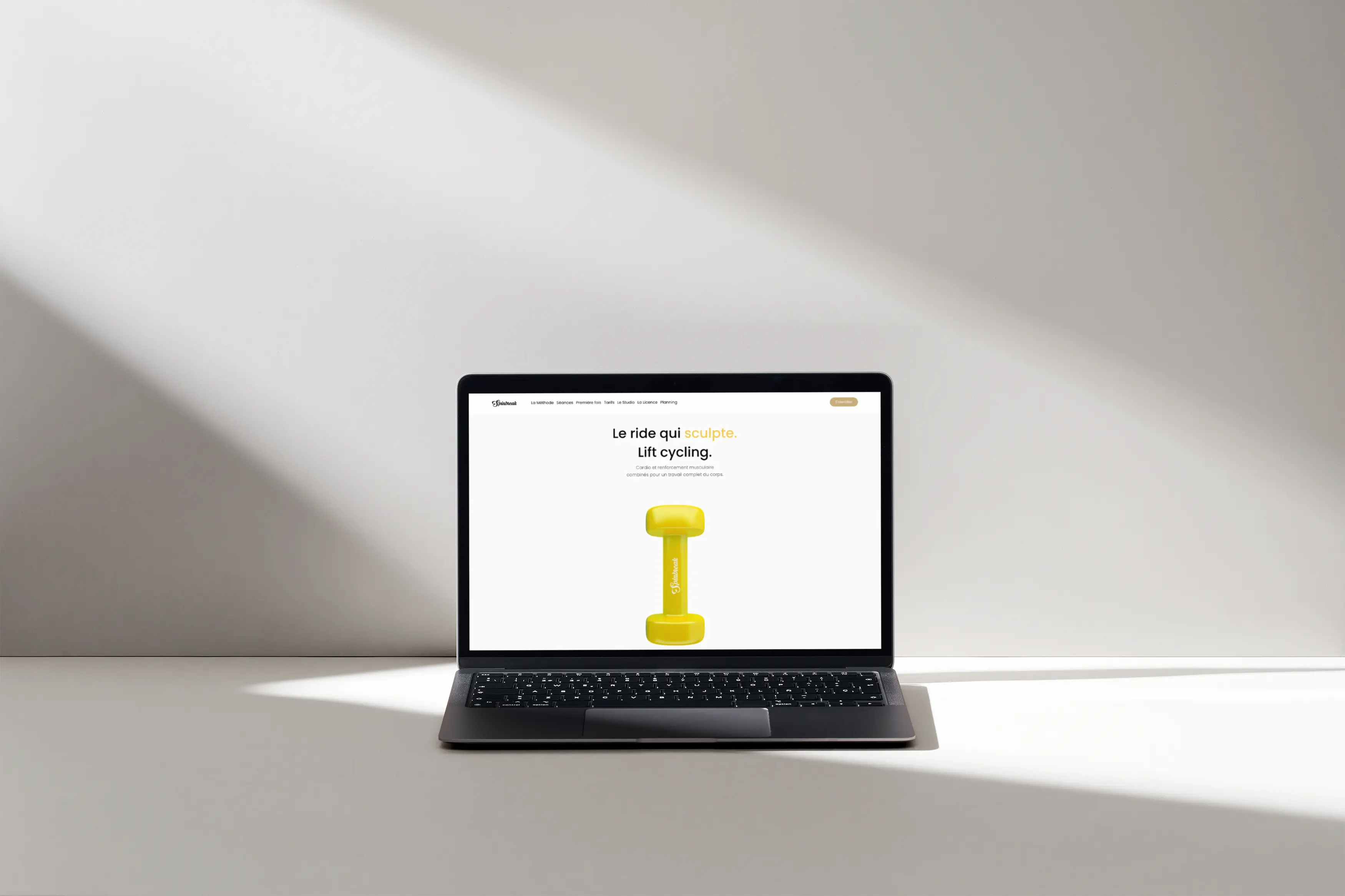 Un ordinateur portable posé affiche une page du site Spinbreak Studio : un visuel épuré avec un haltère jaune au centre et un slogan sur le cycling et le renforcement musculaire.