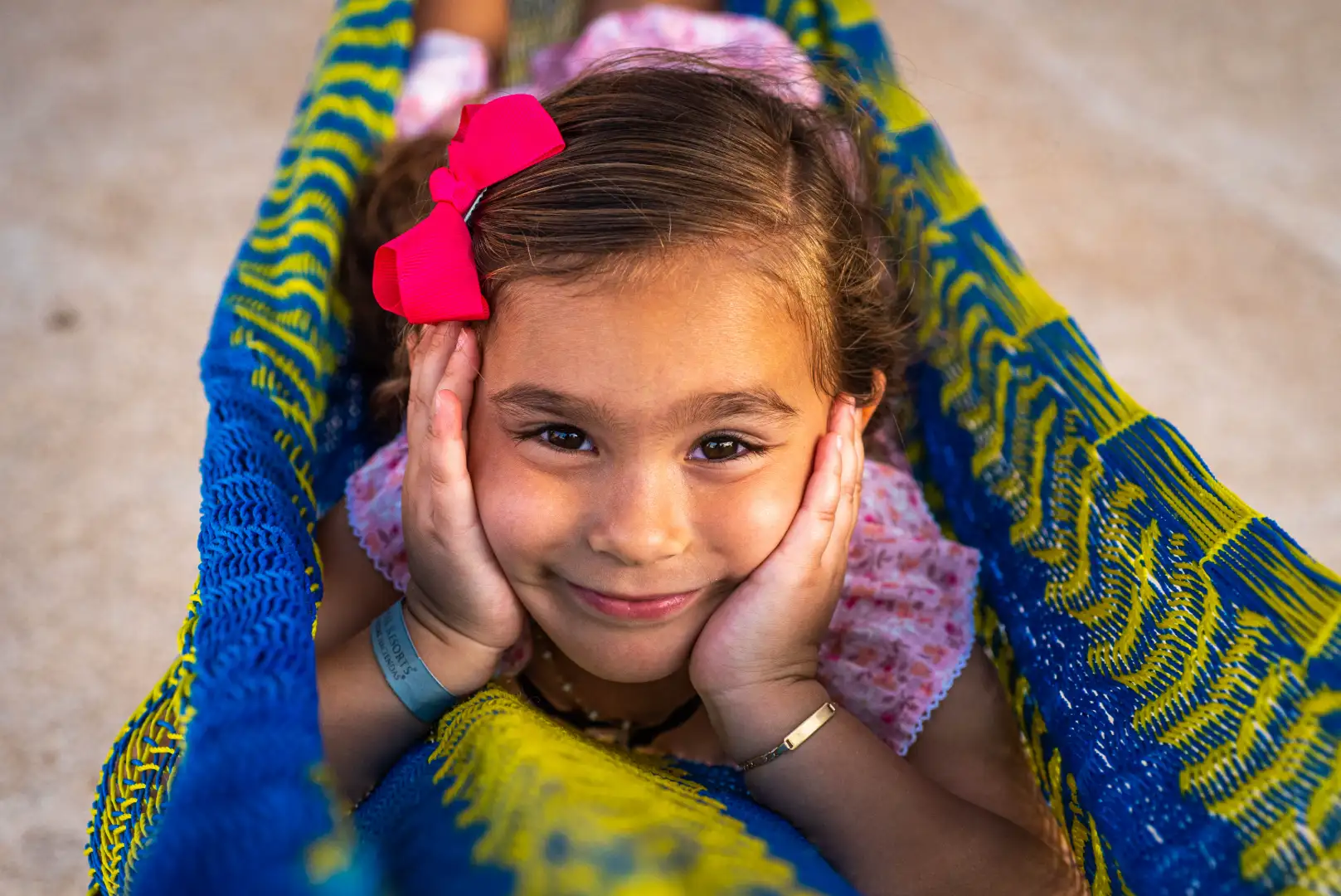 Smiling girl with a pink bow, hands on cheeks, in a blue and yellow hammock.