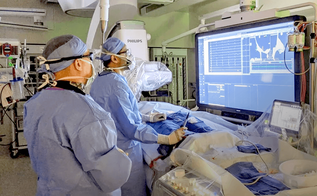 Medical team performing a cardiac procedure in an operating room, with surgeons in scrubs working around advanced medical equipment and monitoring screens displaying cardiac data.
