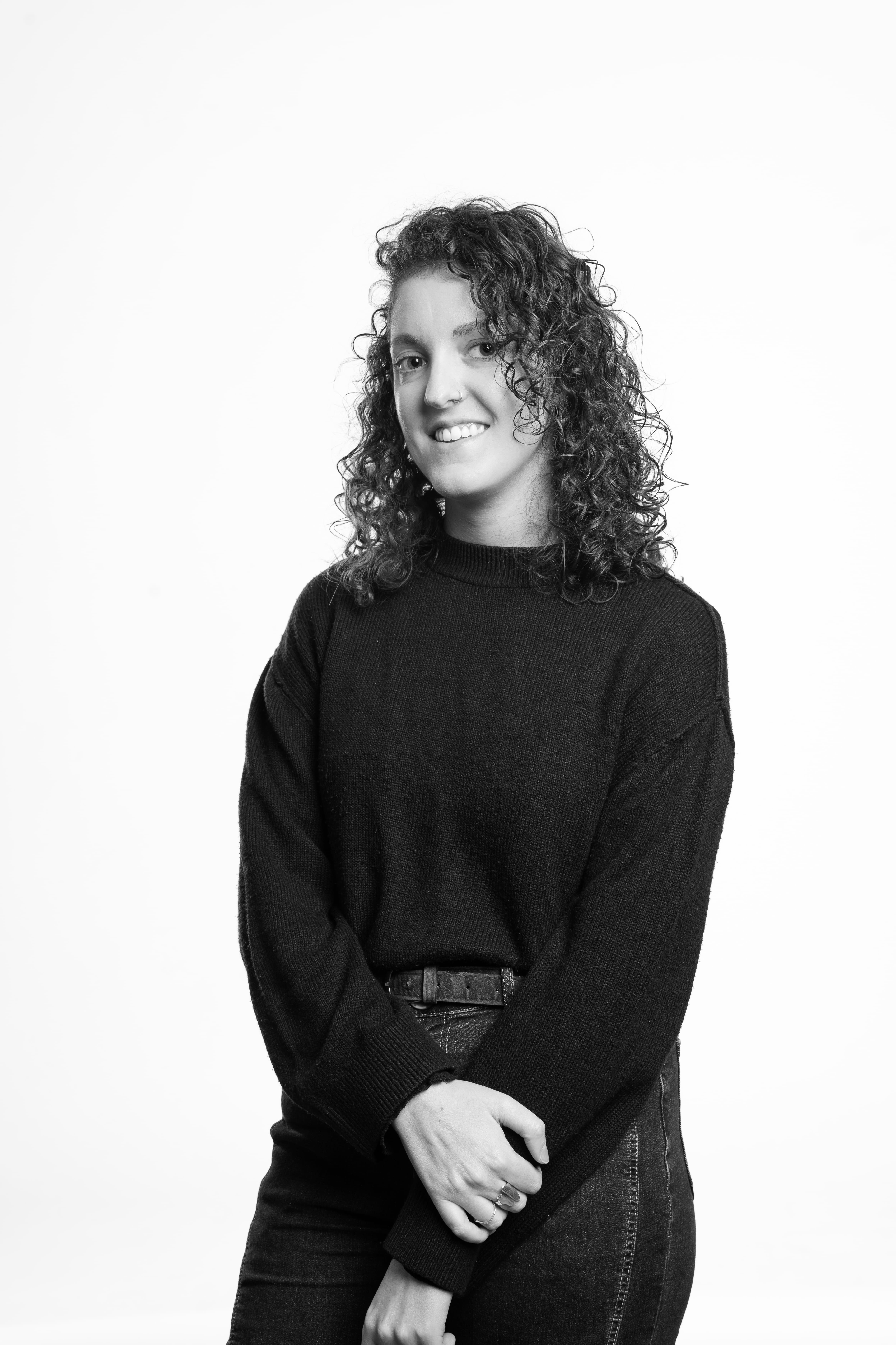 A black and white portrait of a person with curly hair, wearing a dark sweater and jeans. They stand smiling, arms crossed, against a plain background.