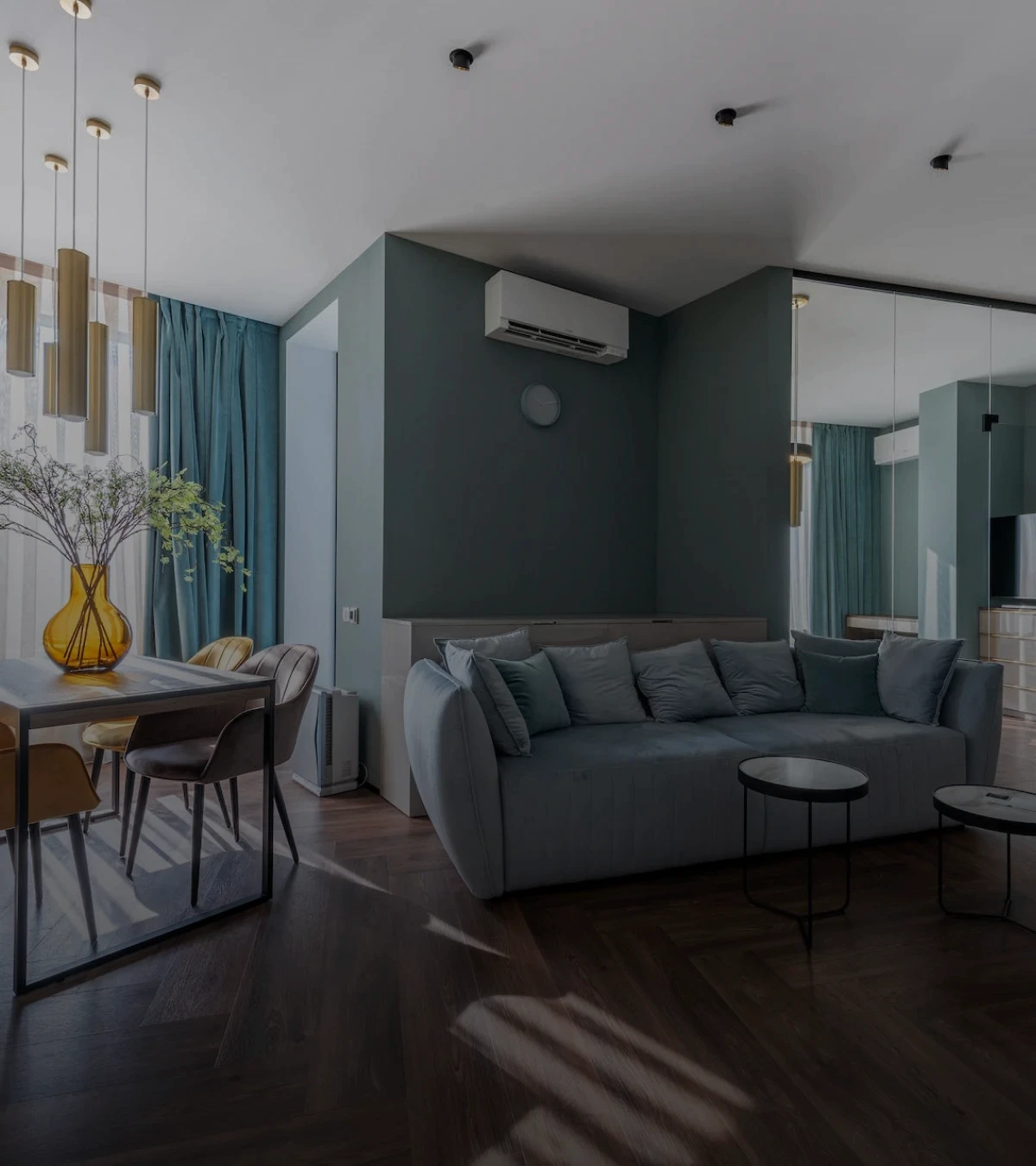 imagem da sala de uma casa elegante projetada por arquitetos. Imagem tem um sombreamento escuro para gerar contraste com o texto da seção. A sala apresenta cores pastéis em tons de azul, cinza e bege.