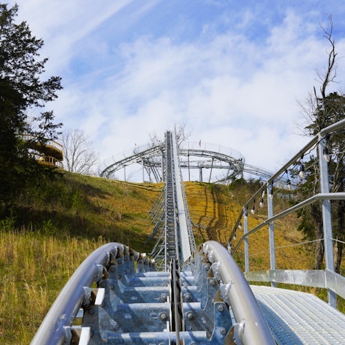 એક ધાતુની રોલર કોસ્ટર ટ્રેક એક ડોંડી ઉપર ઉંચાઈ રહ્યો છે, જે આસપાસના વૃક્ષો અને ઘાસી જમીનથી ઘેરાયેલાં છે અને આંકરું ચાંદલા આકાશ હેઠળ છે.