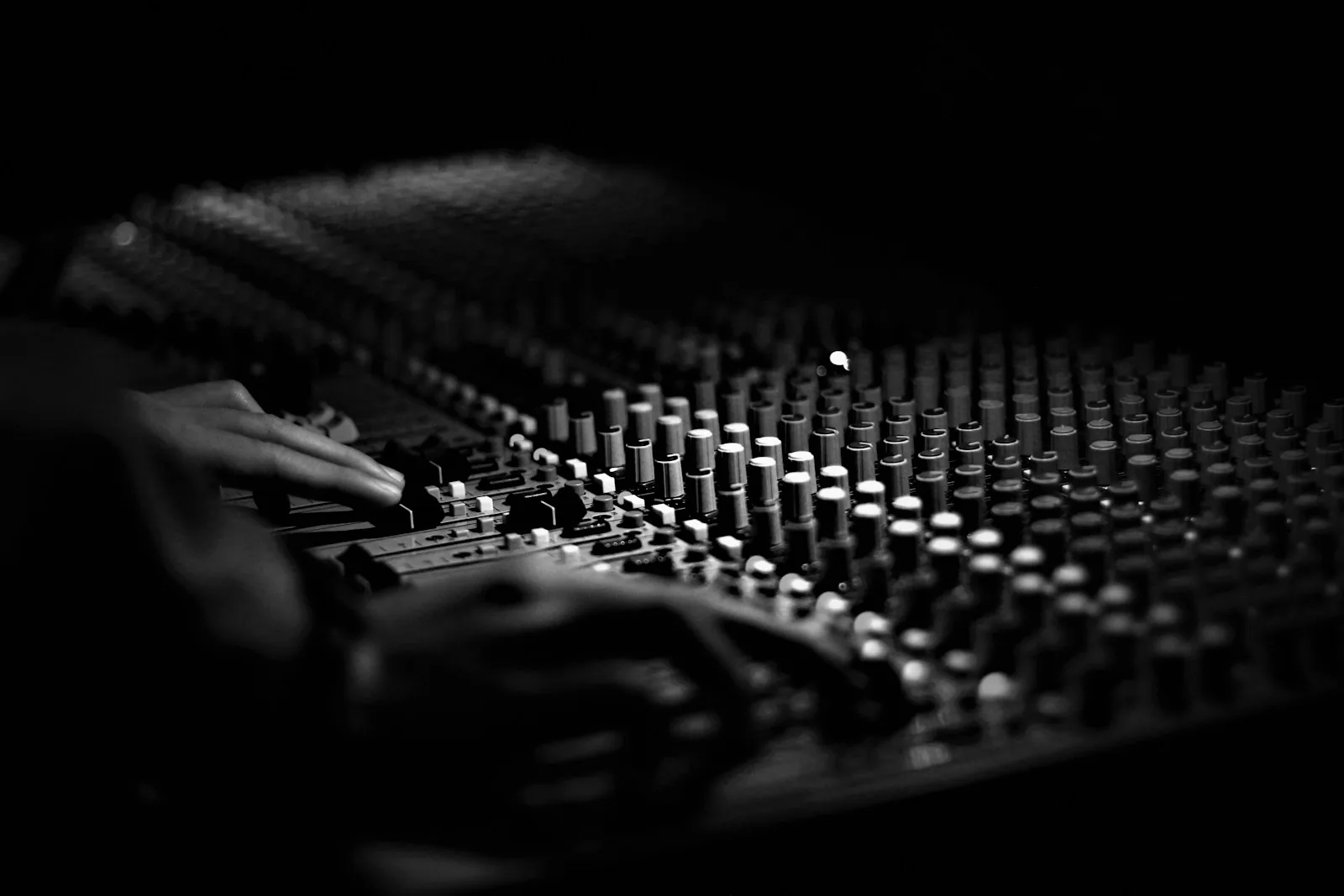 Black and white close-up of two hands moving faders on a large audio mixing console lined with rows of knobs