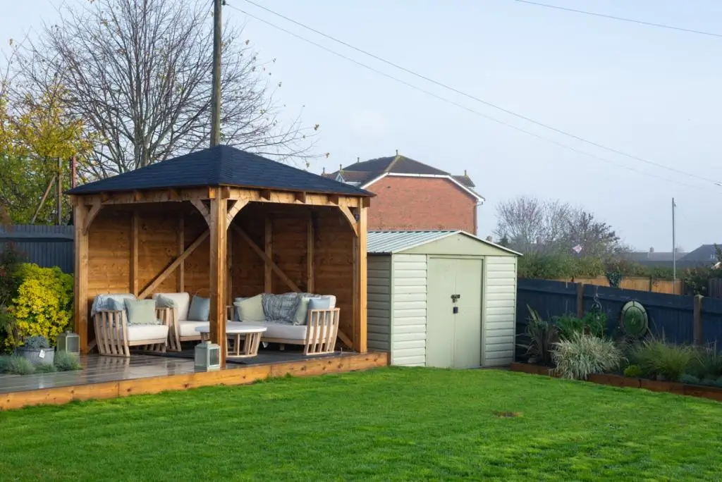 A wooden gazebo with benches stands in a green yard, near a shed and surrounded by trees and a fence.