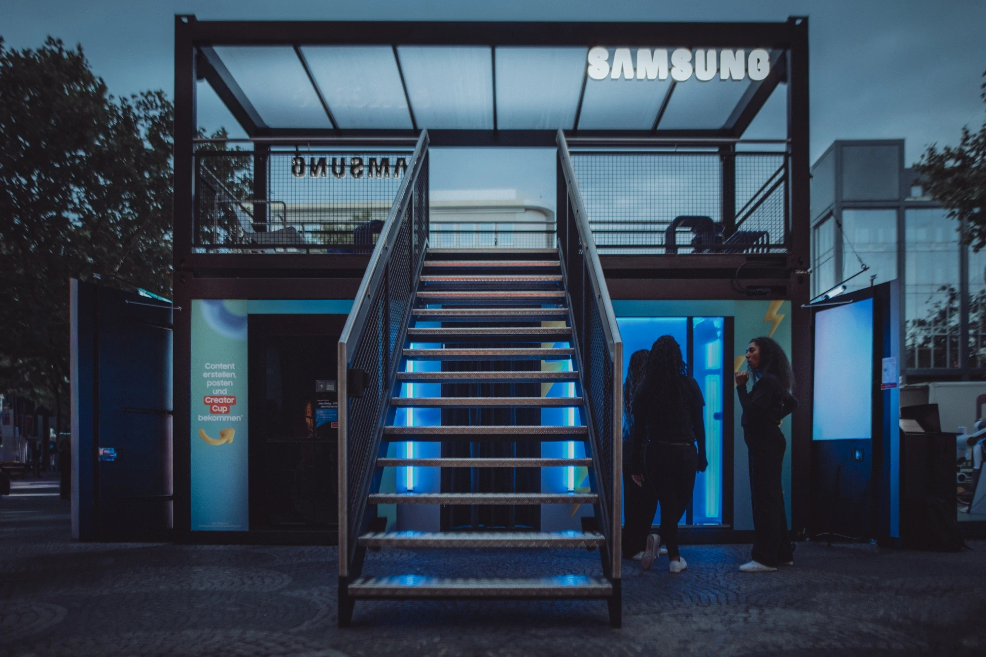 Samsung Creator Cup pop-up booth at dusk, two-storey container structure with steel staircase, illuminated blue interior and Samsung logo on the upper deck.