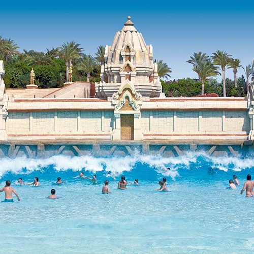 People swimming in a large wave pool with a temple-like structure and palm trees in the background.