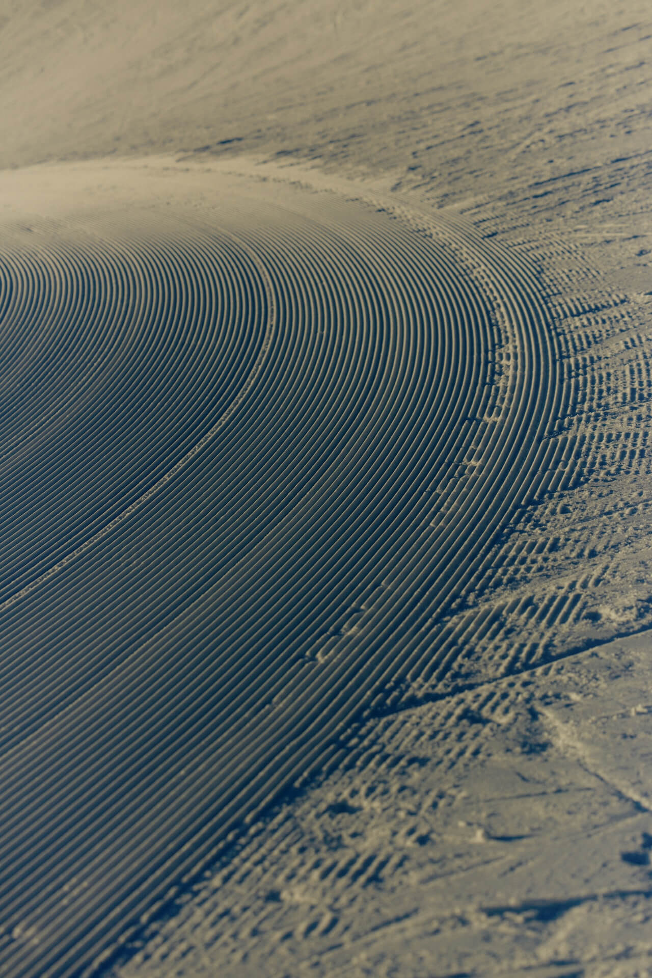 Freshly groomed ski slope with curved lines creating a textured pattern in the snow, evoking a sense of calm and precision in muted lighting.