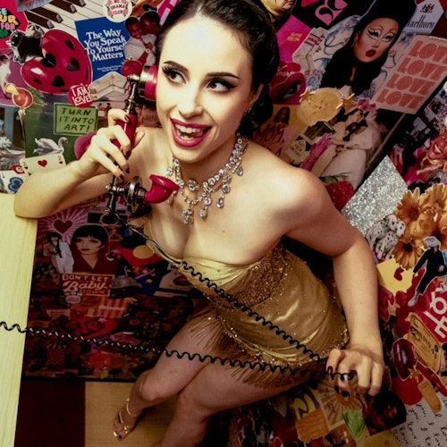 A woman in a gold outfit and necklace smiles while holding a vintage red phone in a booth decorated with colorful posters.