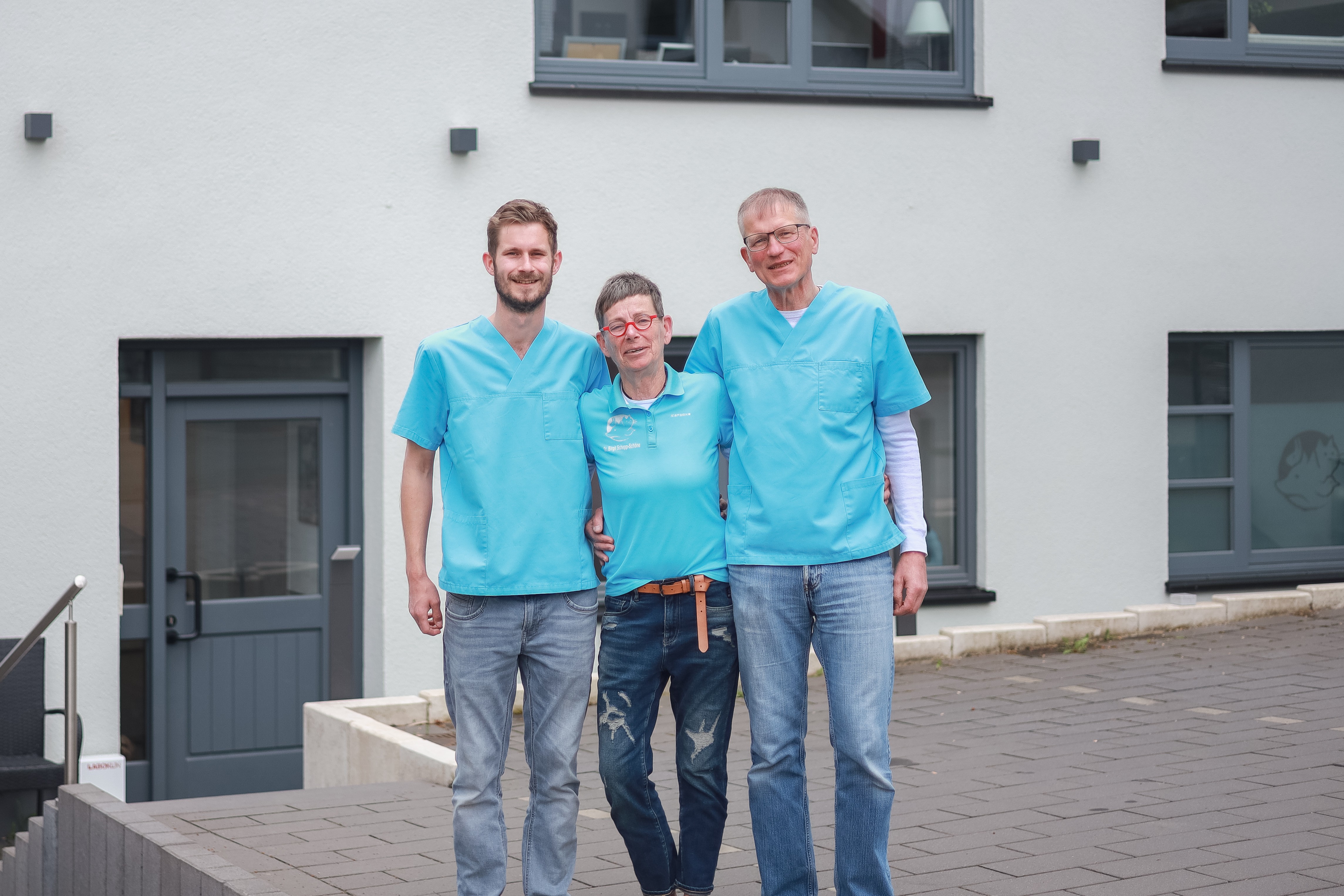 Drei Personen vor Tierarztpraxis. Lächeln in die Kamera, tragen türkisfarbene Oberteile. Tierarzt.