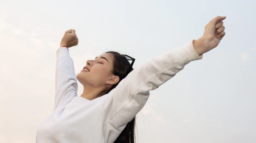 A young woman wearing a white sweatshirt joyfully stretches her arms upwards against a clear sky, exuding a sense of freedom and happiness.