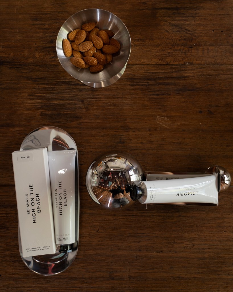 A metal bowl containing almonds sits on a wooden table alongside two tubes of toothpaste displayed on a reflective tray.
