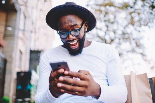 Man with a hat and sunglasses smiling while looking at his phone outdoors.