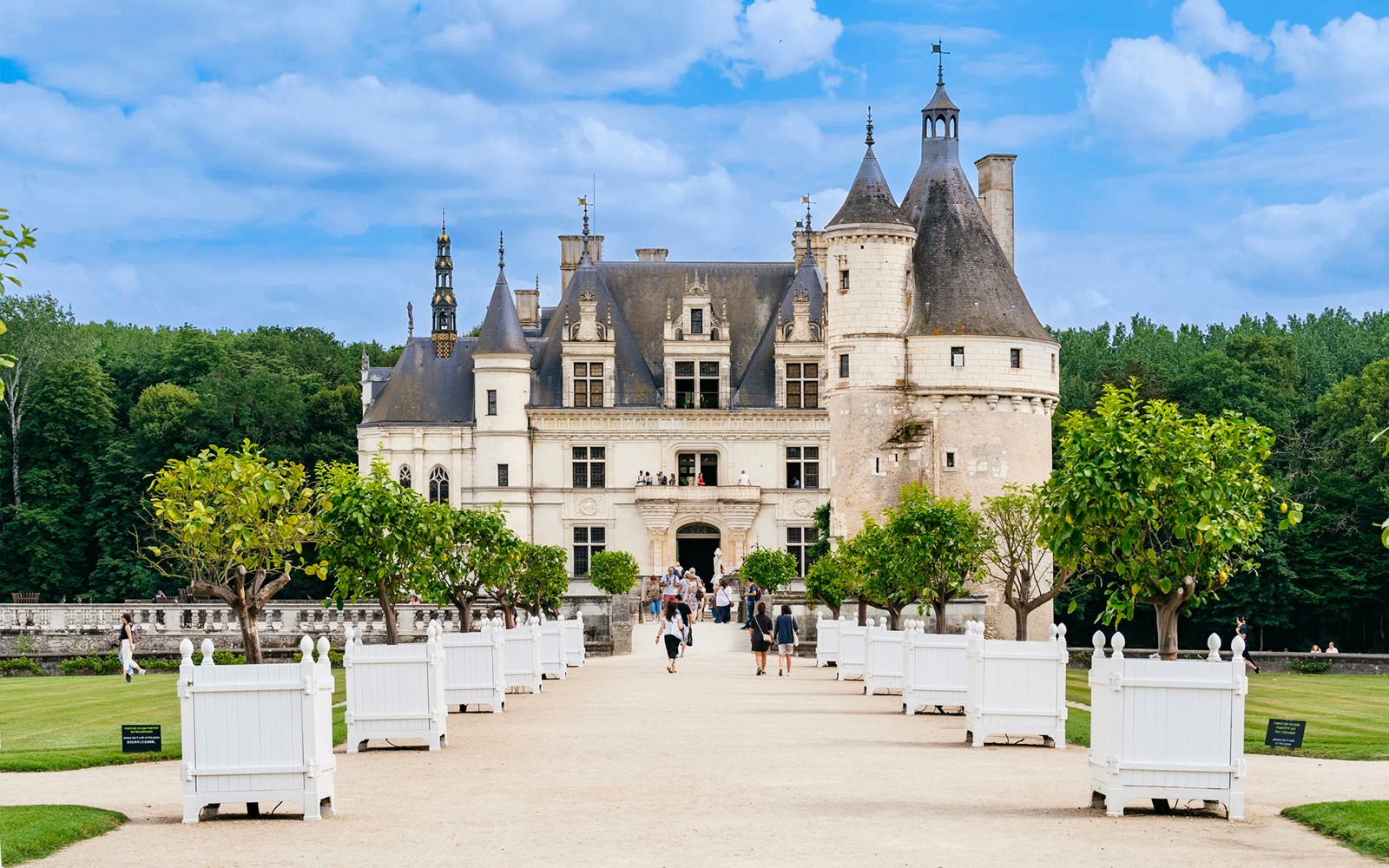 Chenonceau Castle exterior with visitors walking along the tree-lined path.