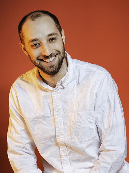 A man with short dark hair and a beard is smiling at the camera. He wears a white long-sleeved button-down shirt against a solid orange-red background. The shirt has visible wrinkles and creases, particularly across the chest and shoulders. His head is slightly tilted to the left, and his gaze is directed toward the camera.