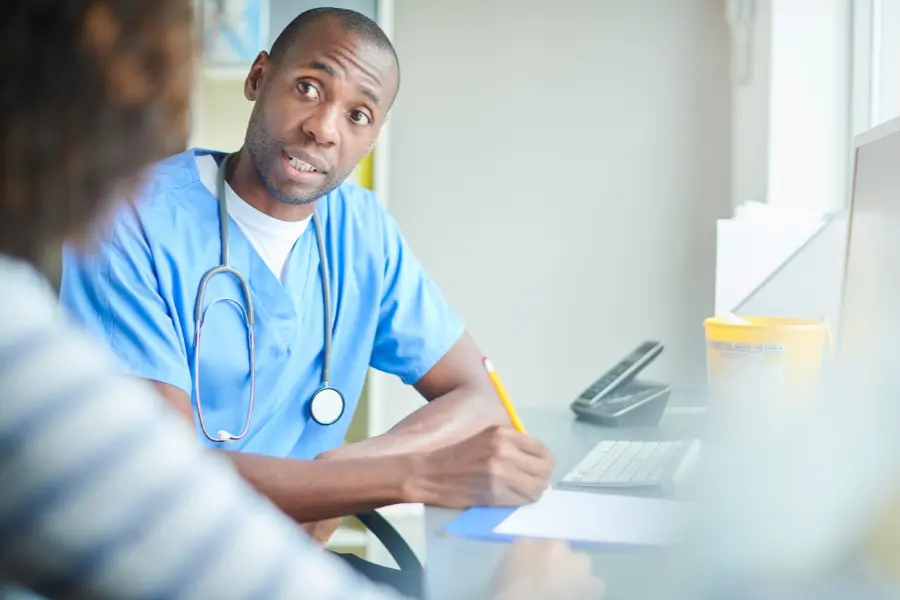 Male nurse in blue scrubs attentively taking notes while consulting a patient using AI medical software at a clinic desk.