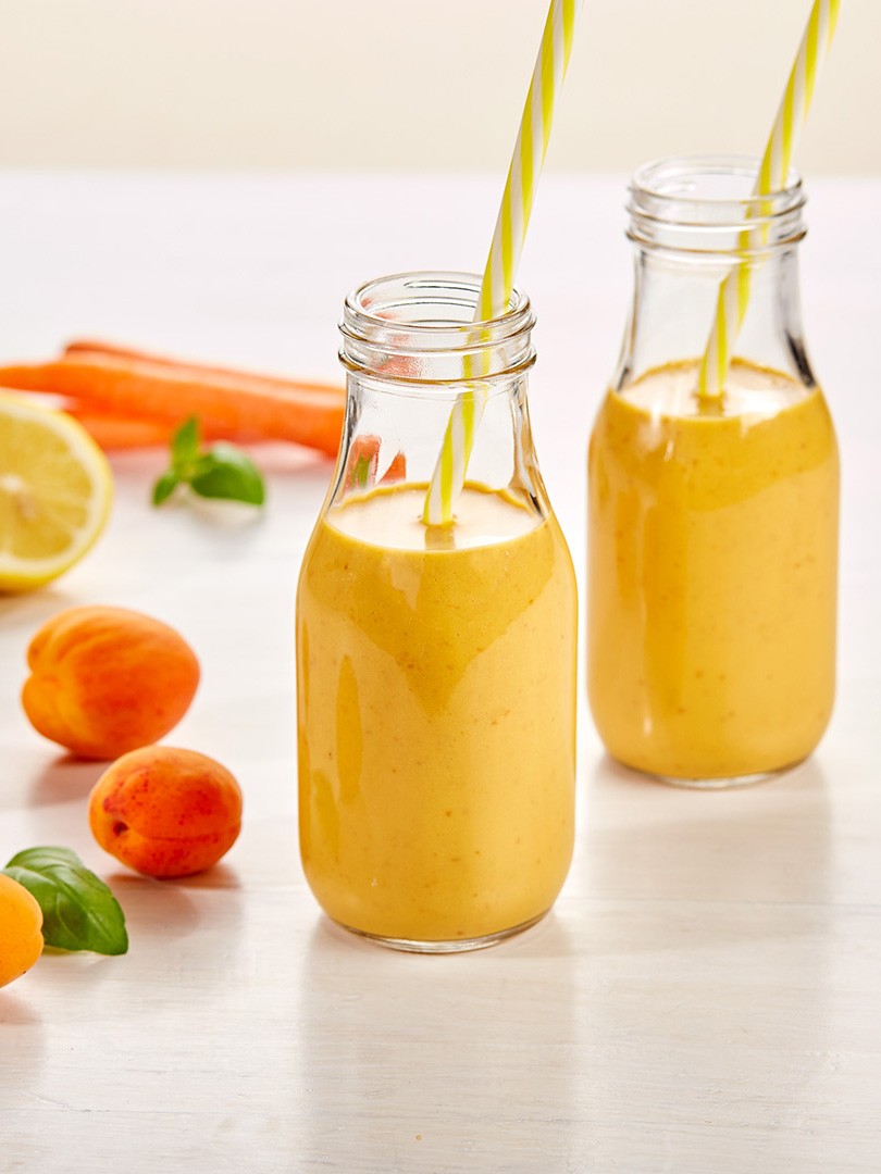 Two glass bottles filled with smoothies are placed on a white table, surrounded by fresh apricots, a sliced lemon, carrots, and basil leaves, each bottle featuring a yellow and white striped straw.
