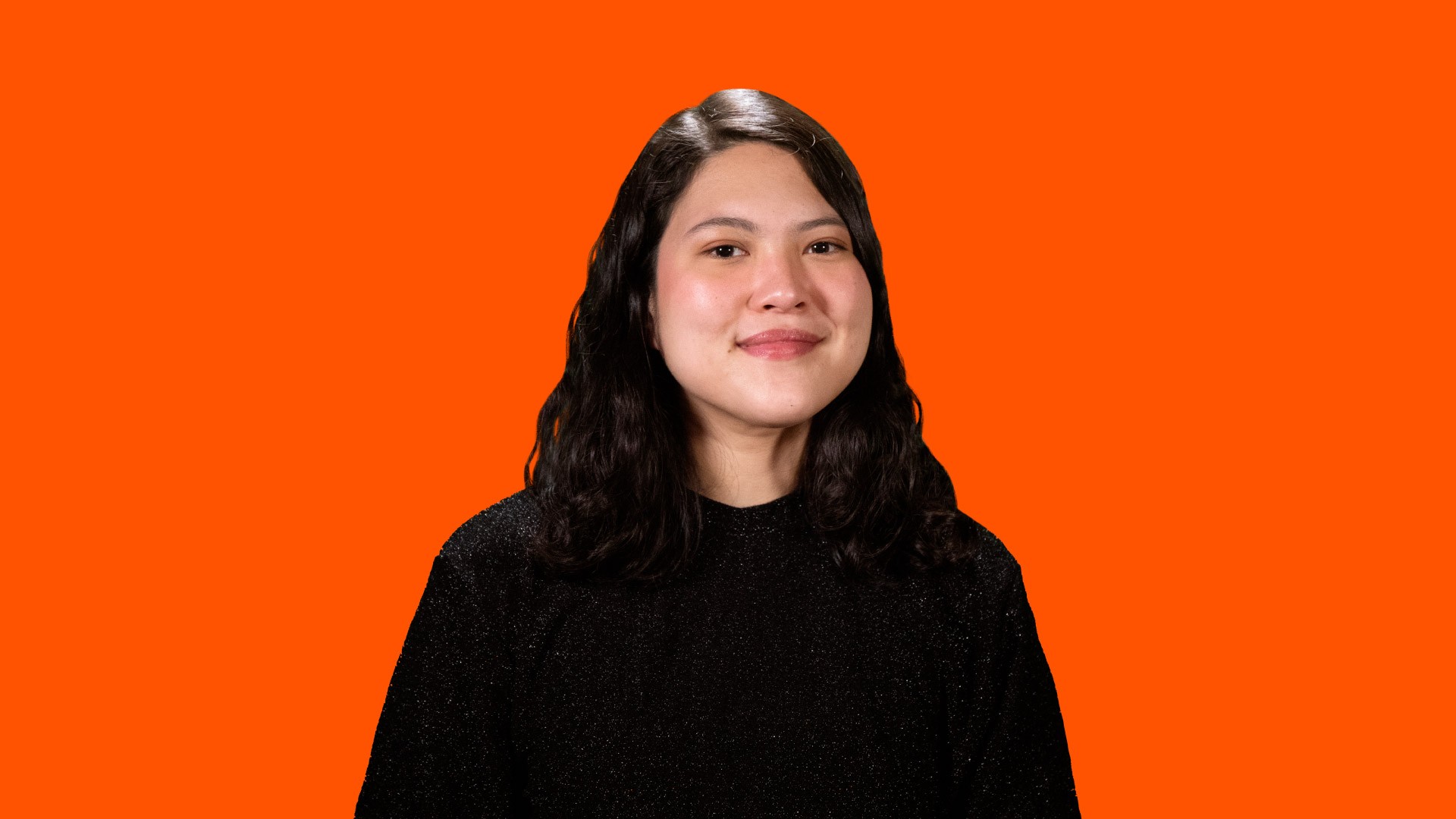 A woman with dark brown hair and light skin stands against a solid orange background. She wears a black top with a subtle textured pattern. Her gaze is directed slightly upward and to the right, and she has a soft smile.