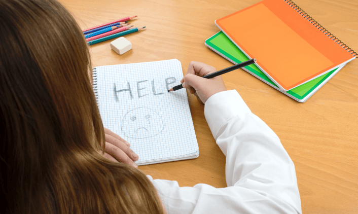 Person writing “HELP” with a sad face in a notebook.