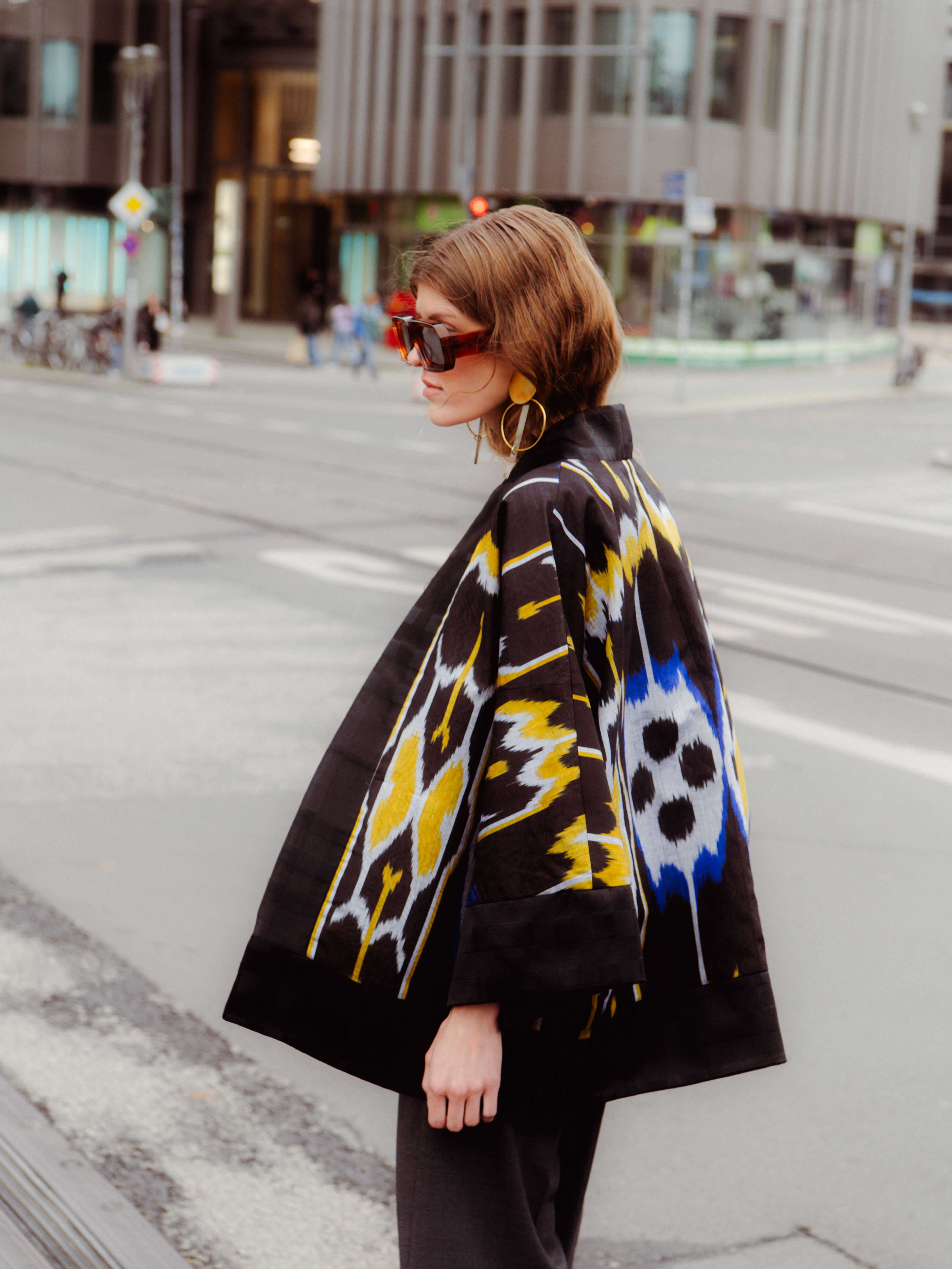 Woman in oversized sunglasses and large earrings walking in an urban setting, wearing a black coat with yellow, white, and blue patterns.