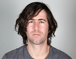 A man with medium-length dark brown hair and light skin is shown in a head-and-shoulders portrait against a plain light gray background. He is wearing a dark gray crew neck t-shirt. His gaze is directed straight at the camera, and he has a neutral expression.