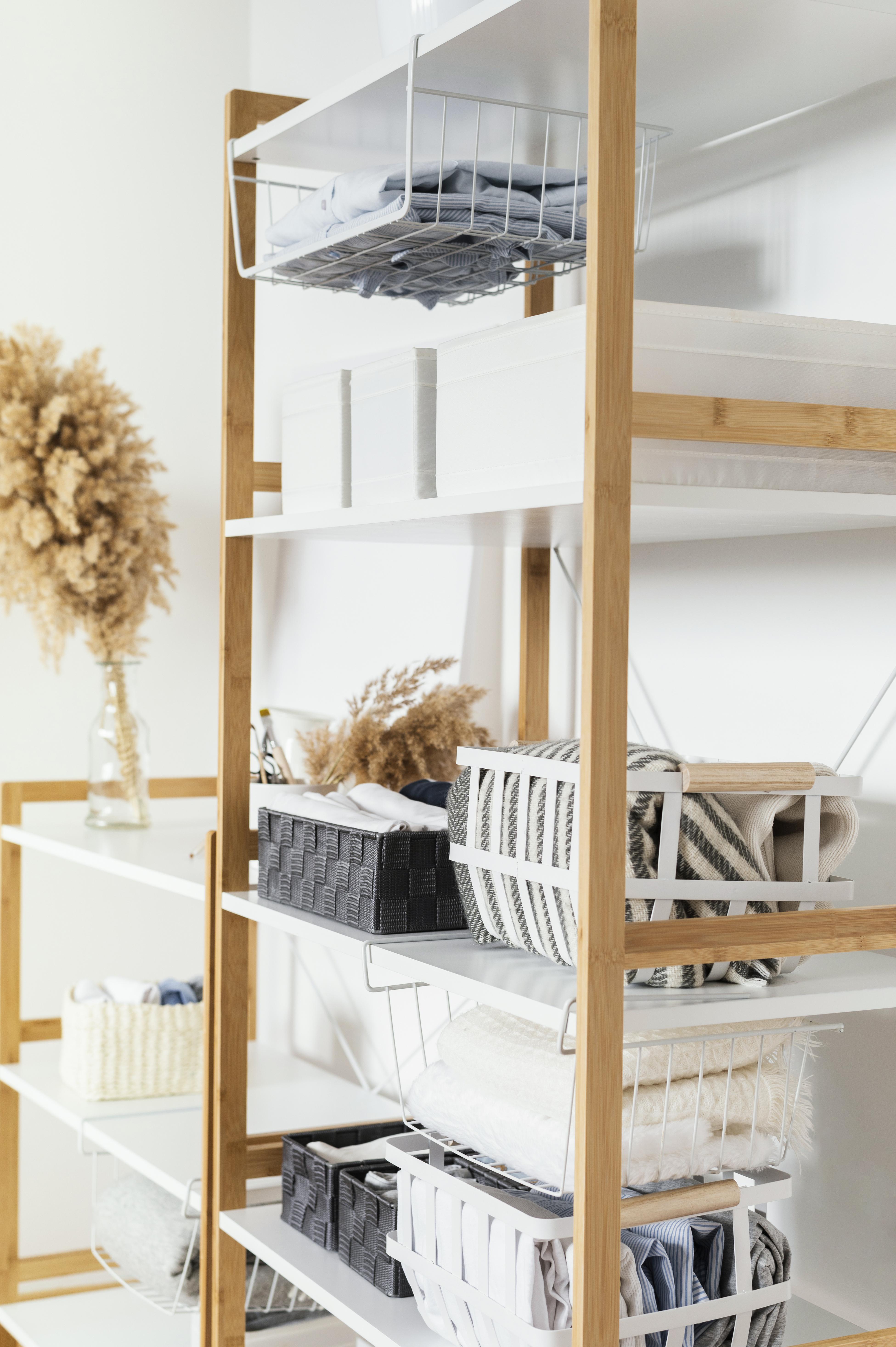A neatly organized bamboo boltless click-in shelving rack holds various woven baskets and folded textiles. A vase with dried pampas grass adds a natural touch, creating a calm, cozy atmosphere.
