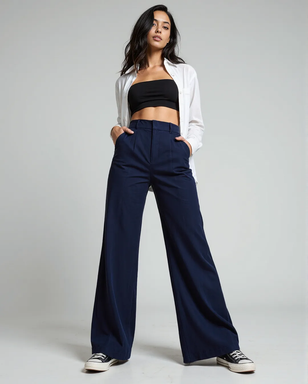 Fashion photograph featuring a minimalist outfit with navy wide-leg trousers, black bandeau top, and white button-up shirt against a neutral gray background.