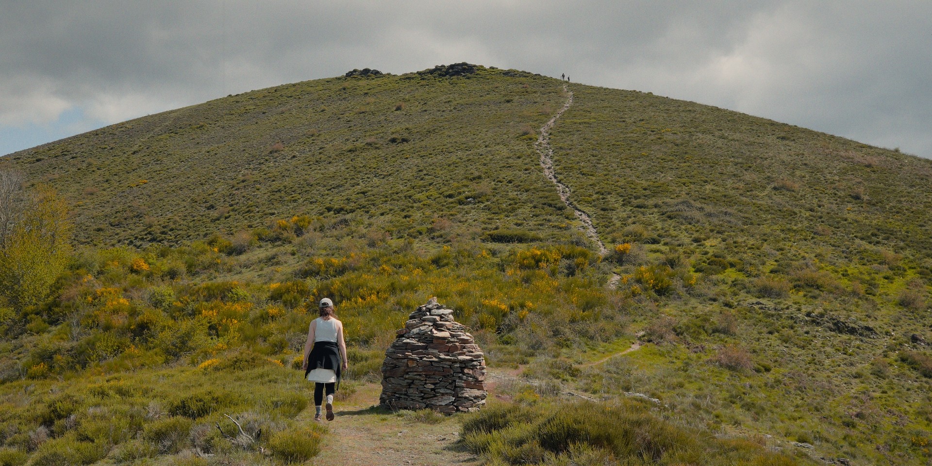 Une femme marche vers un cairn en direction du sommet d'une colline sous un ciel nuageux.