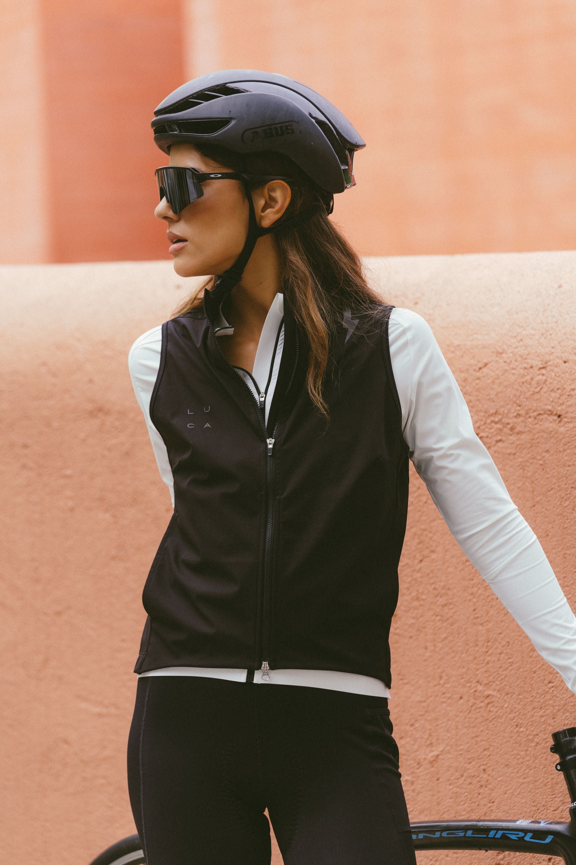 Woman-cyclist-wearing-the-black-Luca-cycling-gilet-fully-zipped-layered-over-a-white-long-sleeve-cycling-jersey-She-is-also-wearing-a-black-helmet-and-large-sunglasses-standing-with-a-bike-against-a-peach-colored-stucco-wall