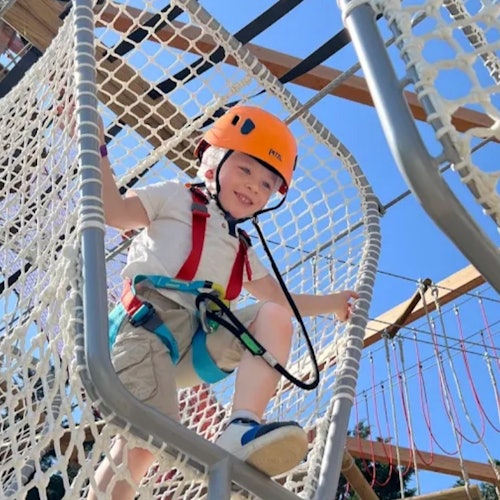 一個戴著頭盔和安全帶的孩子在晴朗的藍天下攀爬一個繩網結構。