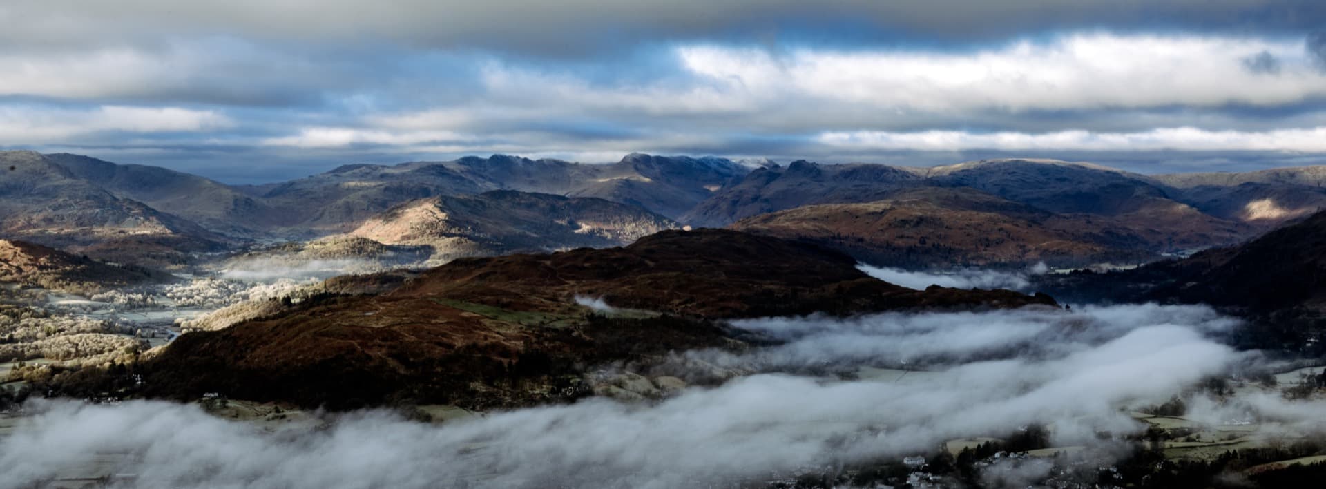 Elevated view from Wansfell Pike showing Lake District fells with low-lying mist filling valleys, layered mountain ridges under dramatic winter sky, sunlit patches on distant peaks