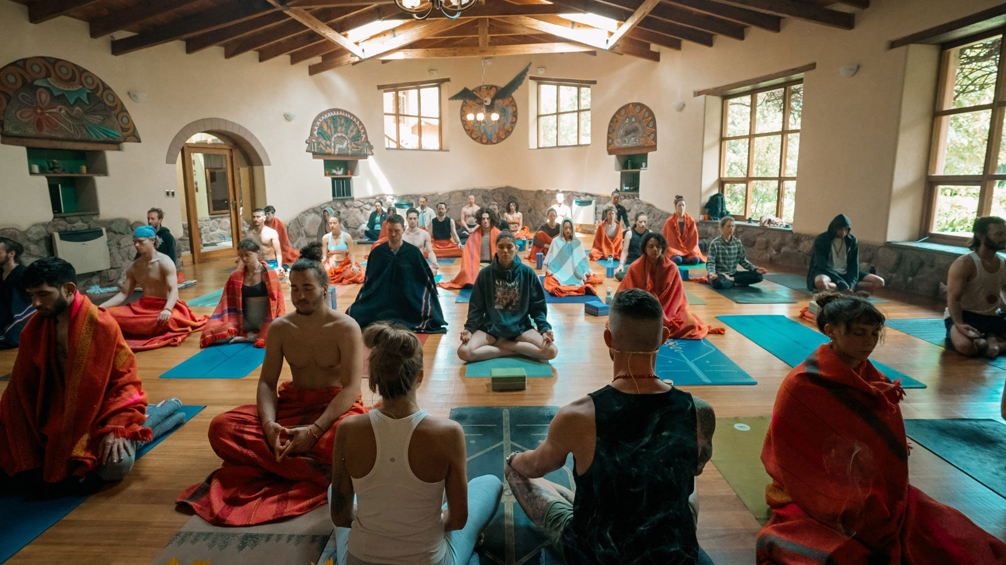 A serene group meditation session in a sunlit room with wooden beams. Participants sit cross-legged on mats, draped in vibrant shawls, conveying tranquility.