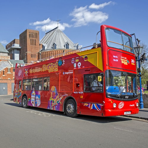 Ein leuchtend roter Doppeldecker-Tourbus mit der Aufschrift "City Sightseeing Stratford", der in einer Straße in der Nähe architektonischer Gebäude geparkt ist.