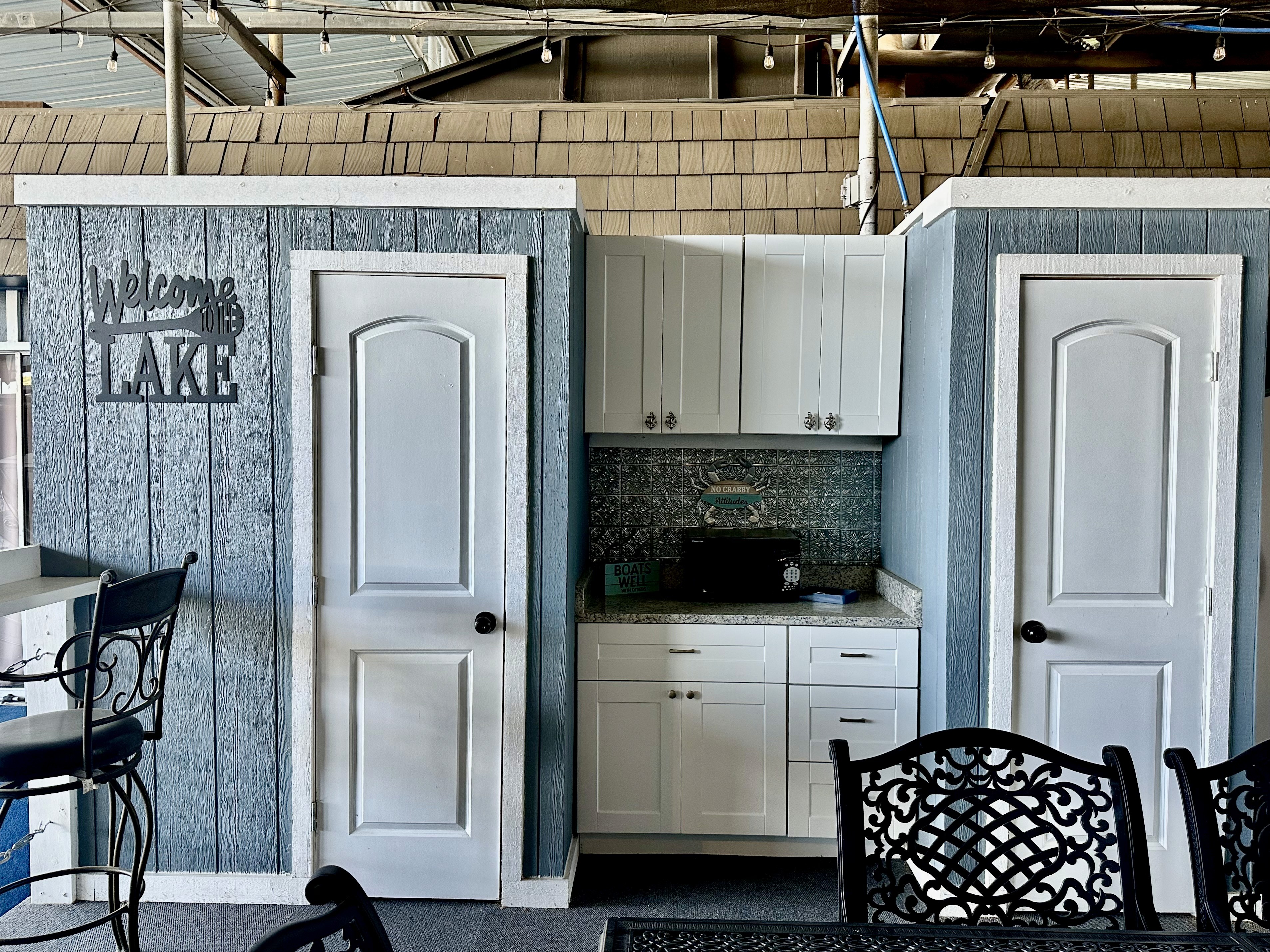 A cozy indoor setup features two white doors on either side of a small kitchenette with a granite countertop and a decorative "Welcome Lake" sign on the blue wooden paneling, creating a welcoming lakeside atmosphere.