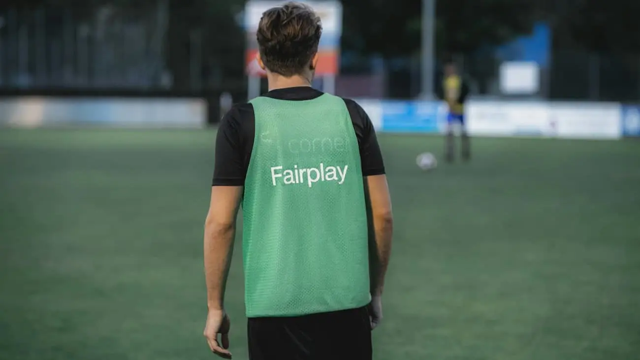 Ein Fußballspieler, der nur von hinten erkennbar ist, trägt eine grüne Trainingsweste mit der Aufschrift "Fairplay". Er steht auf einem Fußballplatz, während andere Spieler und Tore im verschwommenen Hintergrund zu sehen sind. Das Bild verkörpert die Bedeutung von Sportsgeist und dem Geist von Fair Play im Sport. Es ist tagsüber und das klare Wetter betont eine positive und sportliche Umgebung. Dieses Bild veranschaulicht visuell die Bedeutung von Fair Play im Sport und betont die zentrale Rolle von Fair Play im Fußball sowie in anderen Sportarten.