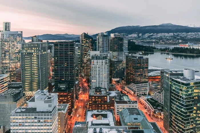 Downtown Vancouver skyline at dusk with glass towers, glowing streets, harbour, and North Shore mountains.