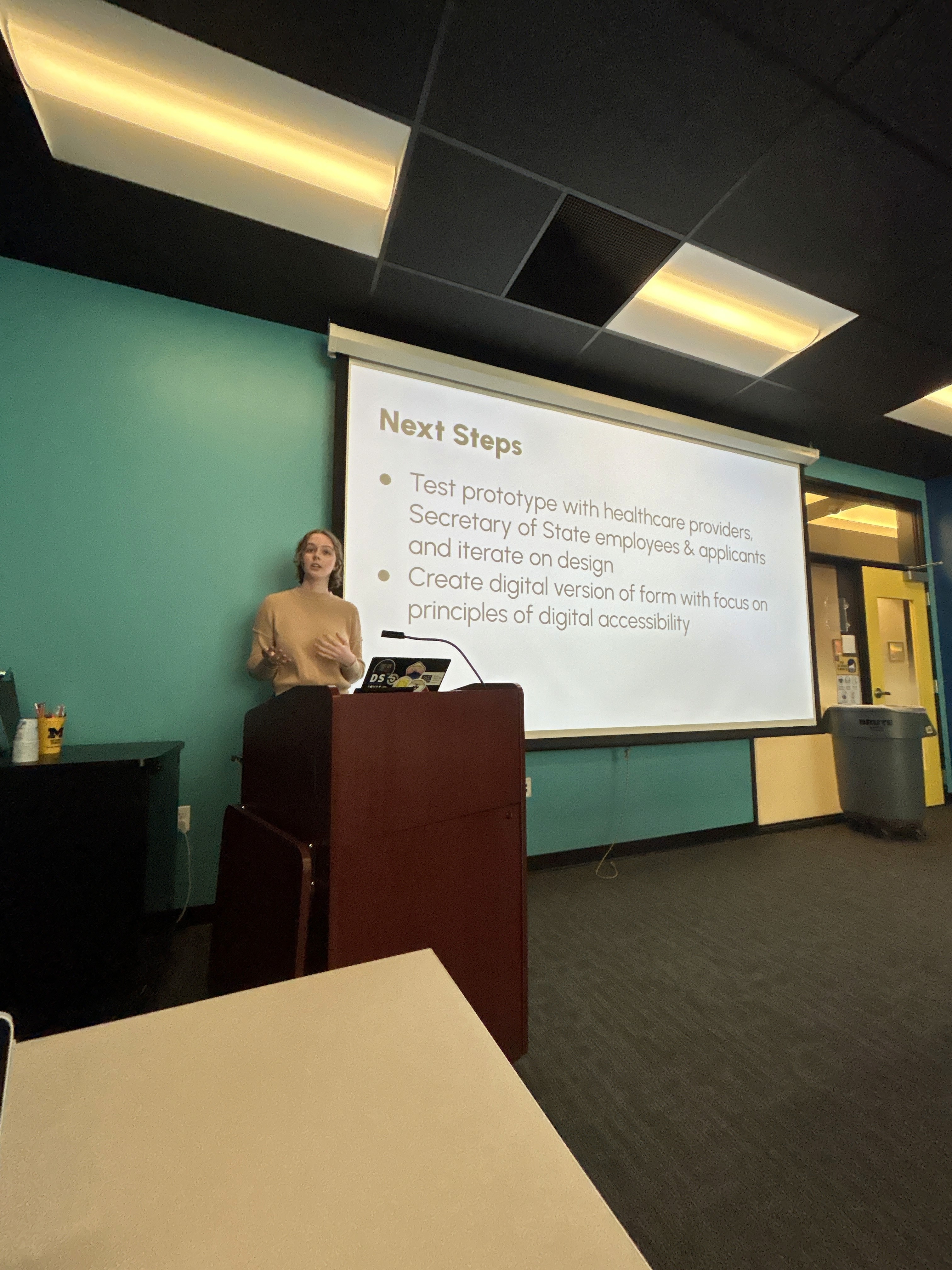 Maddie House presenting at podium. Text on screen behind her reads "Next Steps: Test prototype with healthcare providers, Secretary of State employees & applicants and iterate on design. Create digital version of form with focus on principles of digital accessibility."