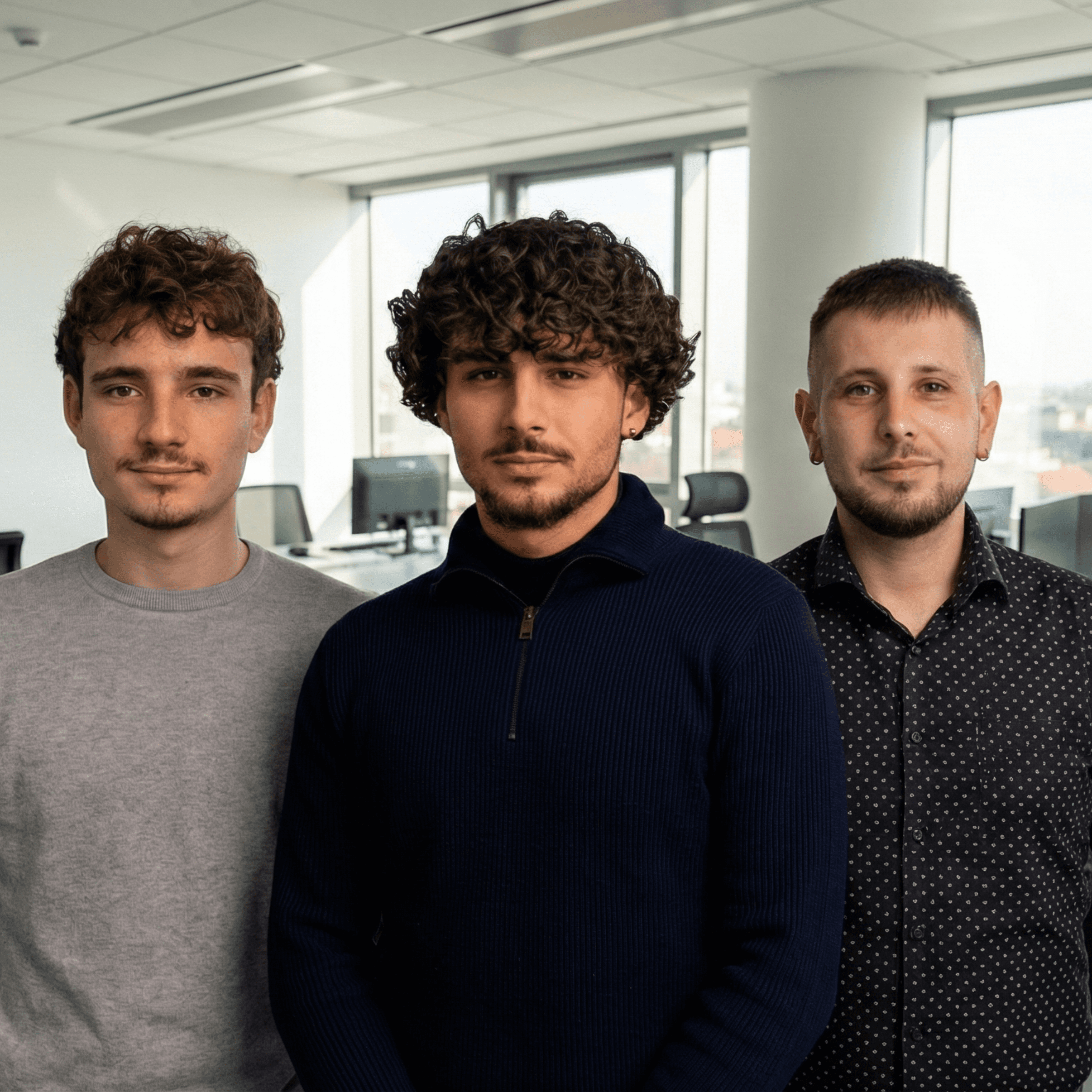 Drei junge Männer stehen zusammen, in Freizeitkleidung, mit einem hellen Büroraum im Hintergrund.