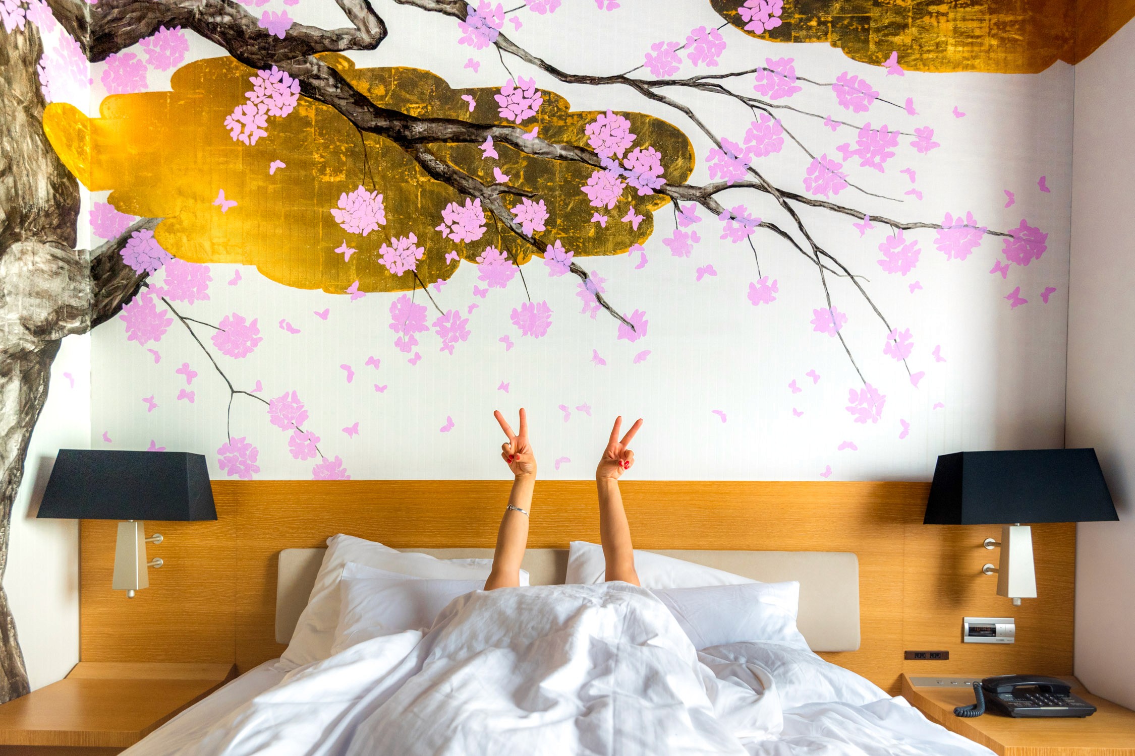 A person lies in a bed with white linens and pillows, legs raised in the air against a mural of pink cherry blossoms.