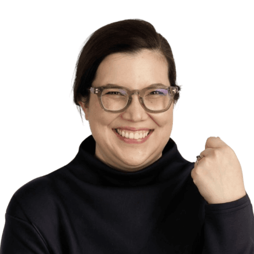 A smiling person with glasses and a dark sweater is raising a fist in a gesture of triumph against a plain background.