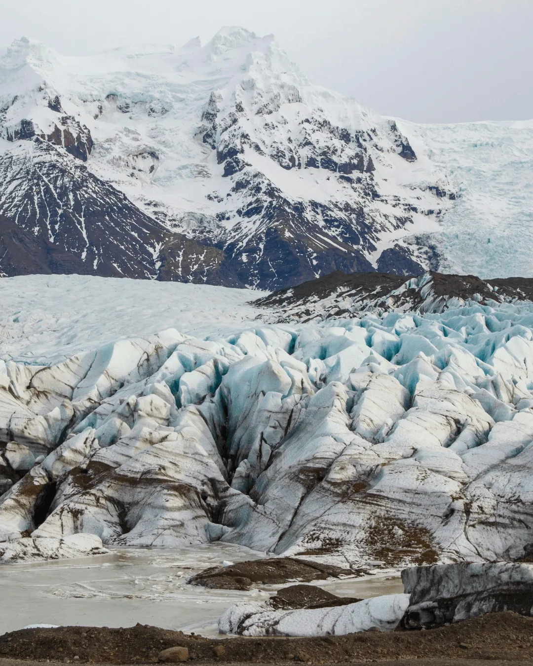 A massive, textured glacier field with deep crevasses and ice ridges under a cloudy Icelandic sky