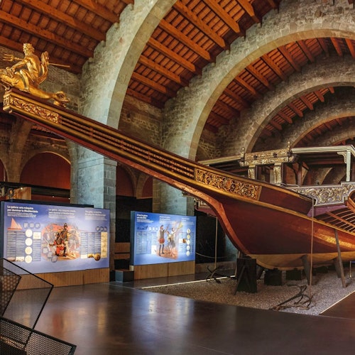 Un gran y ornamentado barco de madera dentro de una exposición de museo con arcos de piedra, con paneles educativos cercanos.