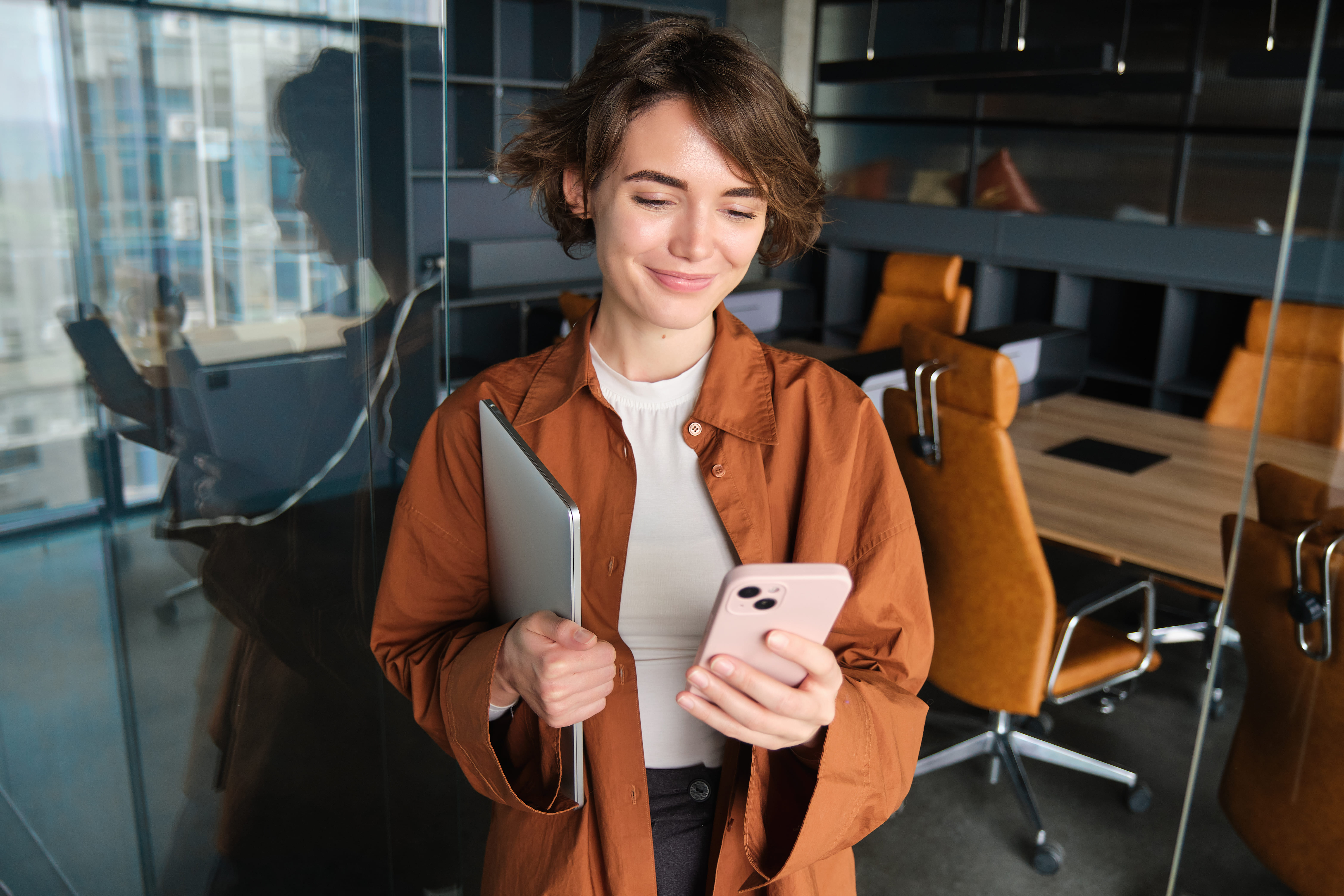 A imagem mostra uma mulher jovem em um ambiente de escritório moderno. Ela tem cabelos curtos e está sorrindo levemente enquanto olha para a tela de um smartphone que segura com uma das mãos. Na outra mão, segura um laptop fechado. Ela veste uma camisa marrom por cima de uma blusa branca. Ao fundo, há uma mesa de reunião com cadeiras de couro laranja e prateleiras embutidas na parede. A luz natural entra através de janelas de vidro, que também refletem parte do cenário. A atmosfera é profissional e descontraída.